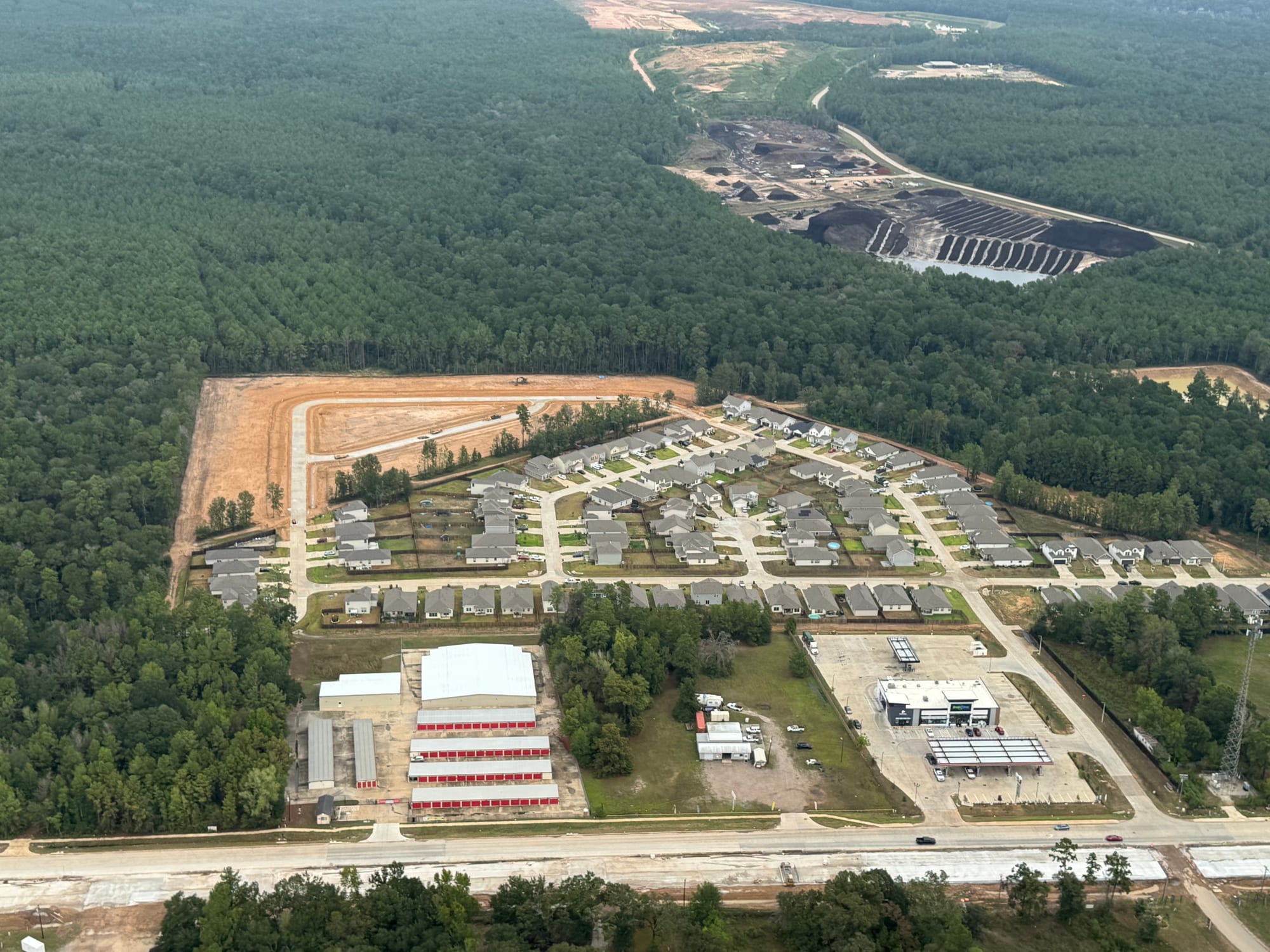 Aerial view of Cedar Crossing: streets of completed single-family homes beside a newly graded section of bare earth with fresh road curbs and a dozer working, bounded by dense forest
