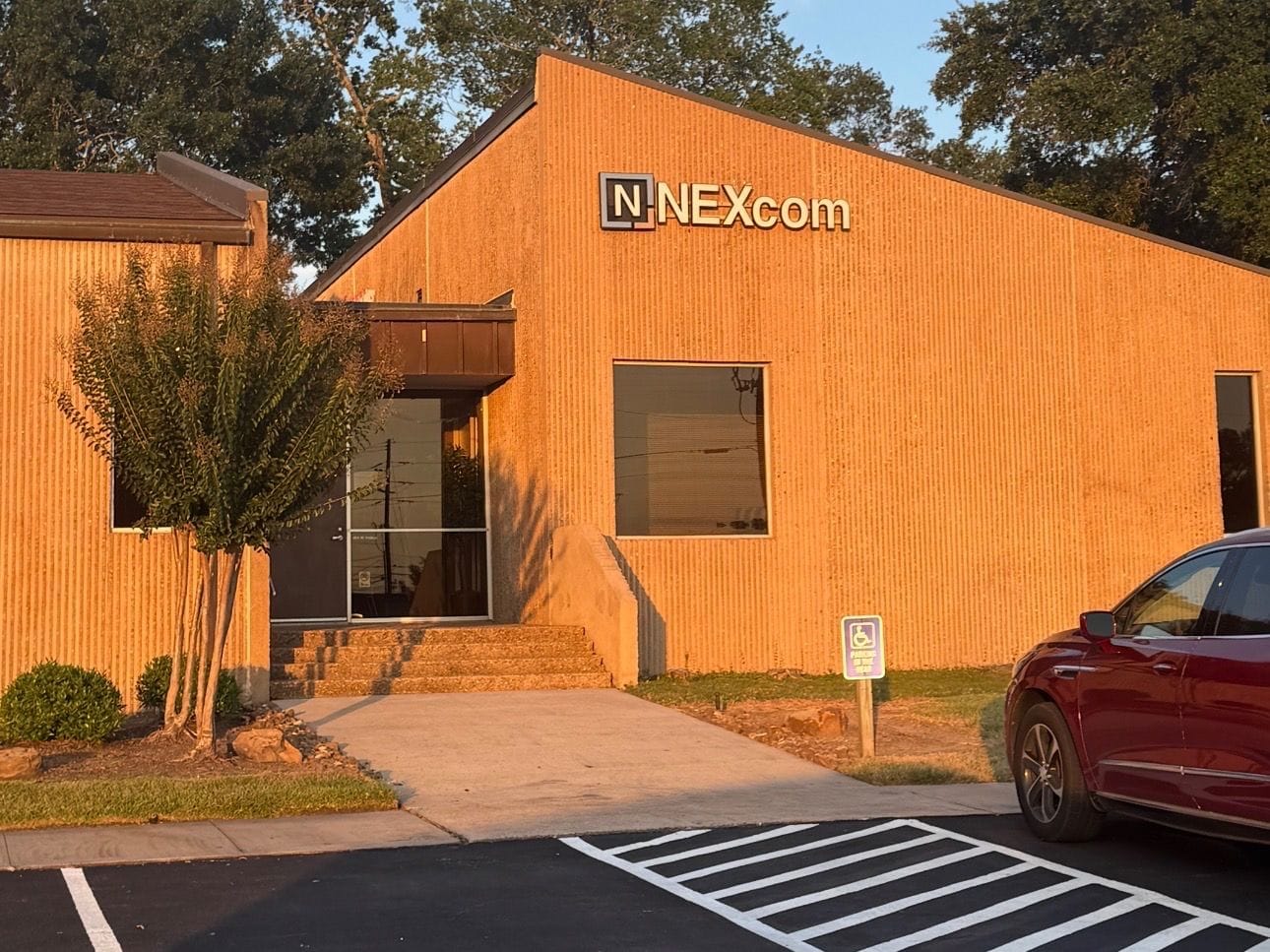 A single-storey orange-brick office building with a NEXcom sign above the entrance, trees around it and a car parked in front