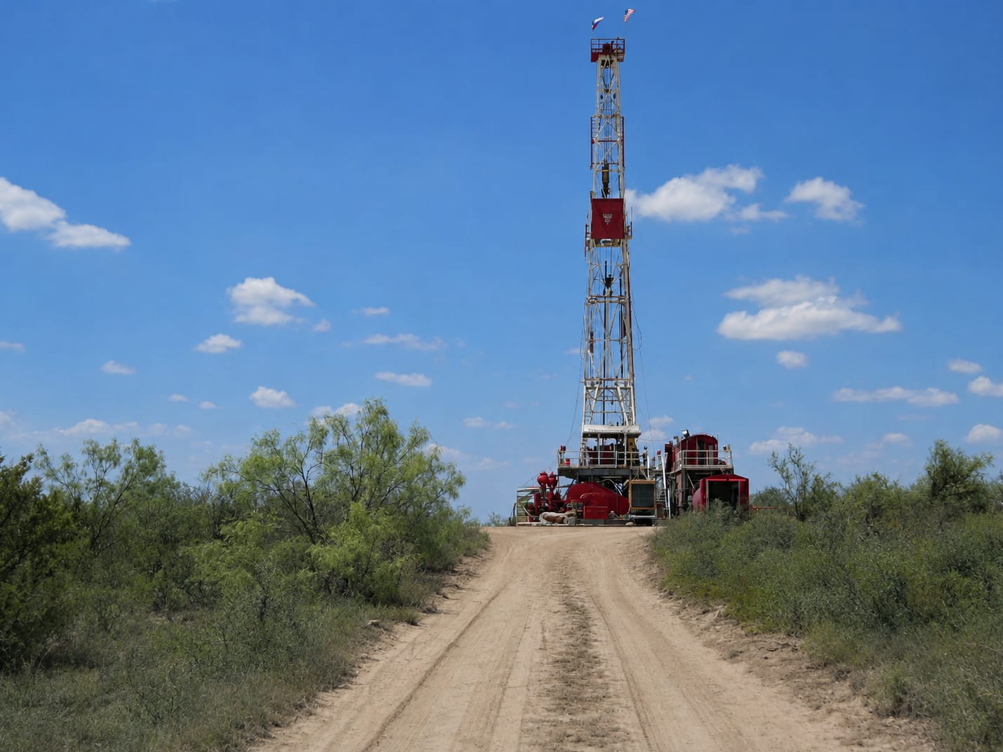 A drilling rig on a dirt road through West Texas scrub, its red and white derrick against a blue sky