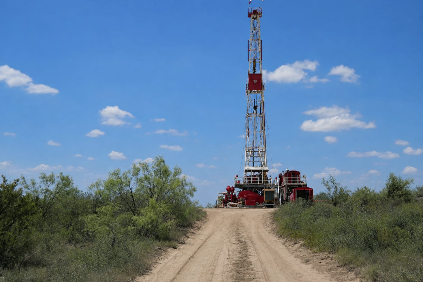 A drilling rig on a dirt road through West Texas scrub, its red and white derrick against a blue sky