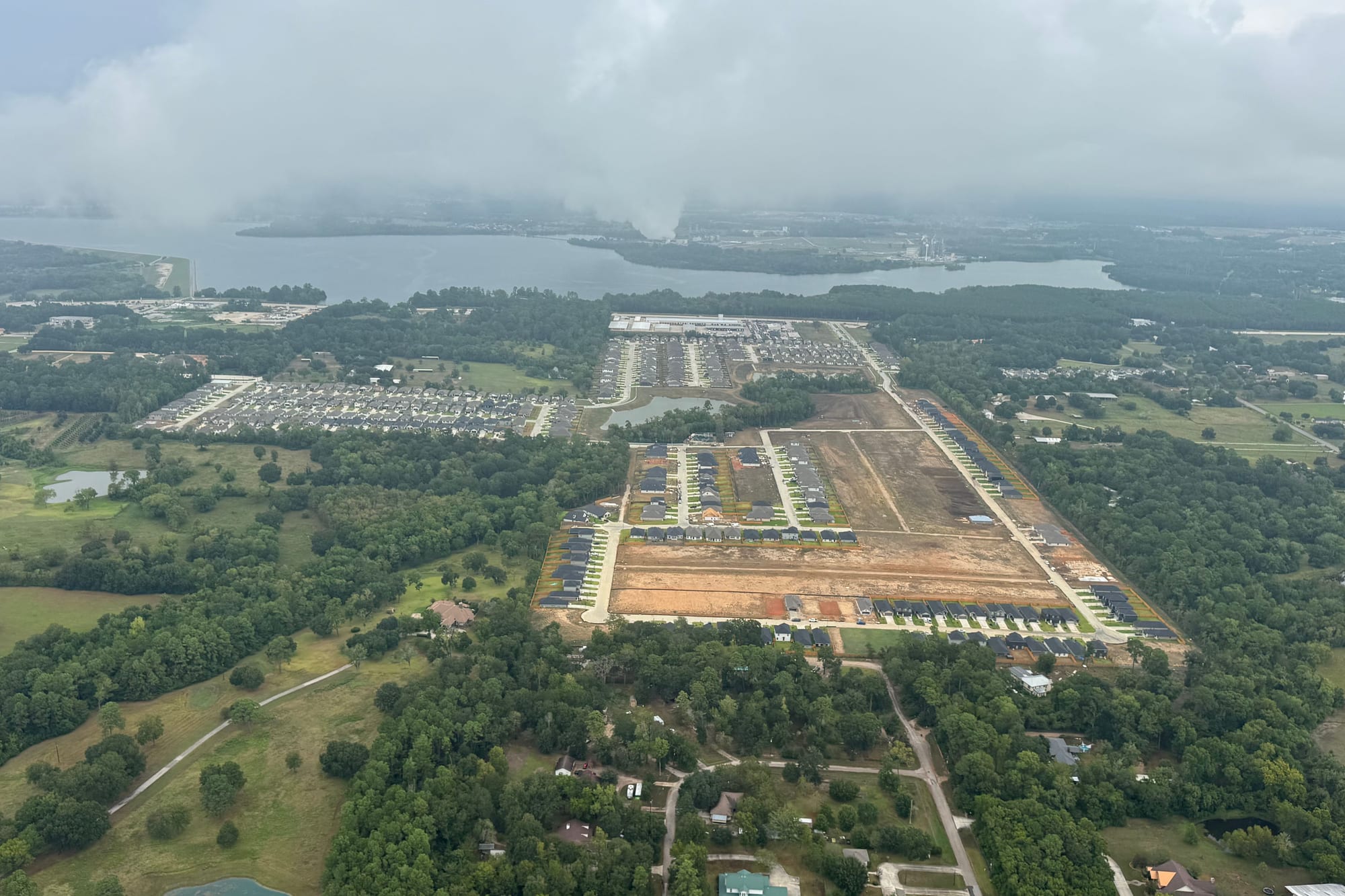 Aerial view of Lexington Heights: streets of completed single-family homes beside a large area of graded bare lots with roads and kerbs already laid, an older subdivision to one side, forest around, and Lake Conroe on the horizon